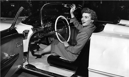 Unidentified woman waves while posing behind the wheel of the Cadillac La Espada concept convertible, at the Cadillac exhibit area on the show floor. Car door is open, giving a partial view of the custom interior, including nacelle headrests for each occupant.